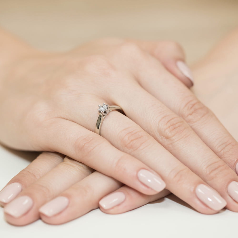 W0222.B3.DIW.08_4 Na dłoniach z delikatnym manicure znajduje się srebrny pierścionek z diamentem.
