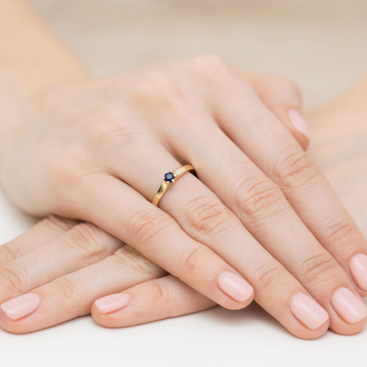 W0222.Z3.SAB.02_8 Złoty pierścionek z niebieskim kamieniem na palcu, dłonie z delikatnym manicure w jasnym kolorze.