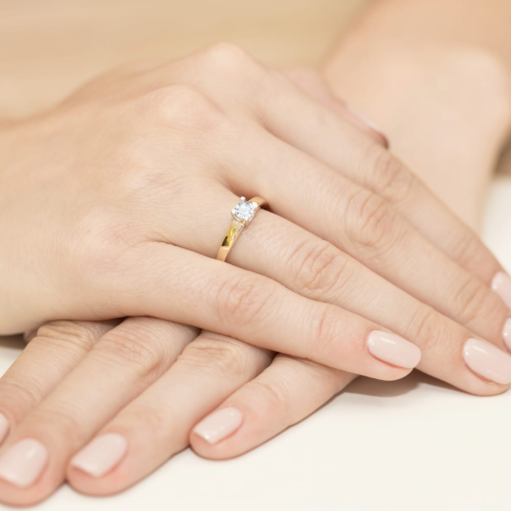 W0225.Z3.DIW.05_4 Dwie dłonie z delikatnym manicure, jedna z pierścionkiem zaręczynowym z diamentem na złotej obrączce.