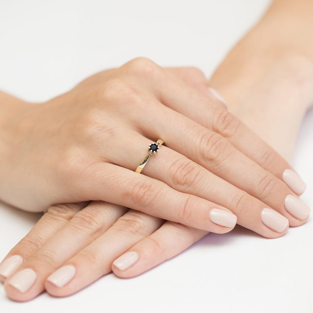 W0228.Z3.DIB.05_4 Złoty pierścionek z czarnym kamieniem na palcu, dłonie z delikatnym manicure w jasnym kolorze.