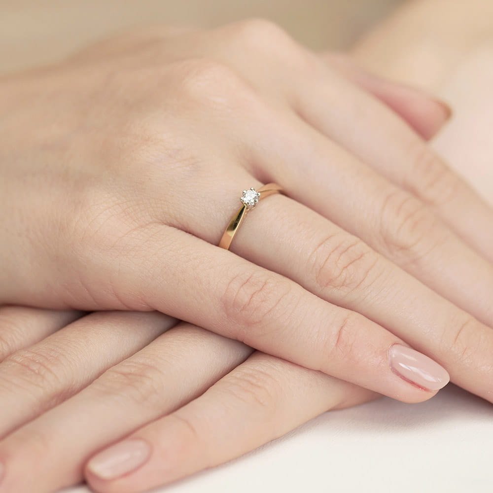 W0229.Z3.LDW.05_4 Złoty pierścionek z diamentem na palcu, otoczony dłońmi z delikatnym manicure.