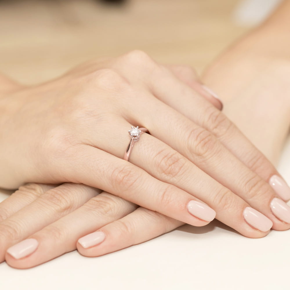 W0234.R3.LDW.04_4 Na dłoni z delikatnym manicure znajduje się pierścionek z diamentem na cienkiej, różowej obrączce.