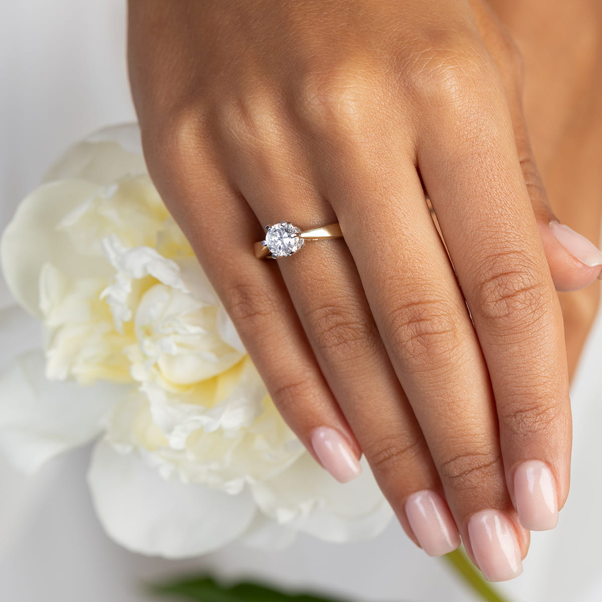 W0234.Z3.LDW.06_4 Złoty pierścionek z dużym diamentem na palcu, trzymanym obok białej piwonii.