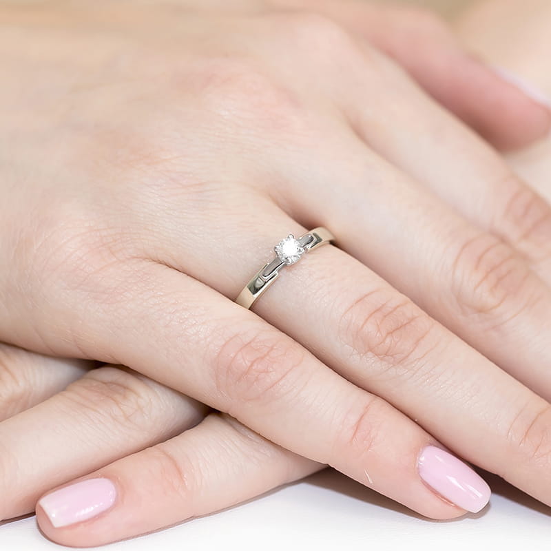 W0241.B3.DIW.03_4 Na dłoni widoczny delikatny pierścionek z małym diamentem, otoczony gładką, srebrną obrączką.