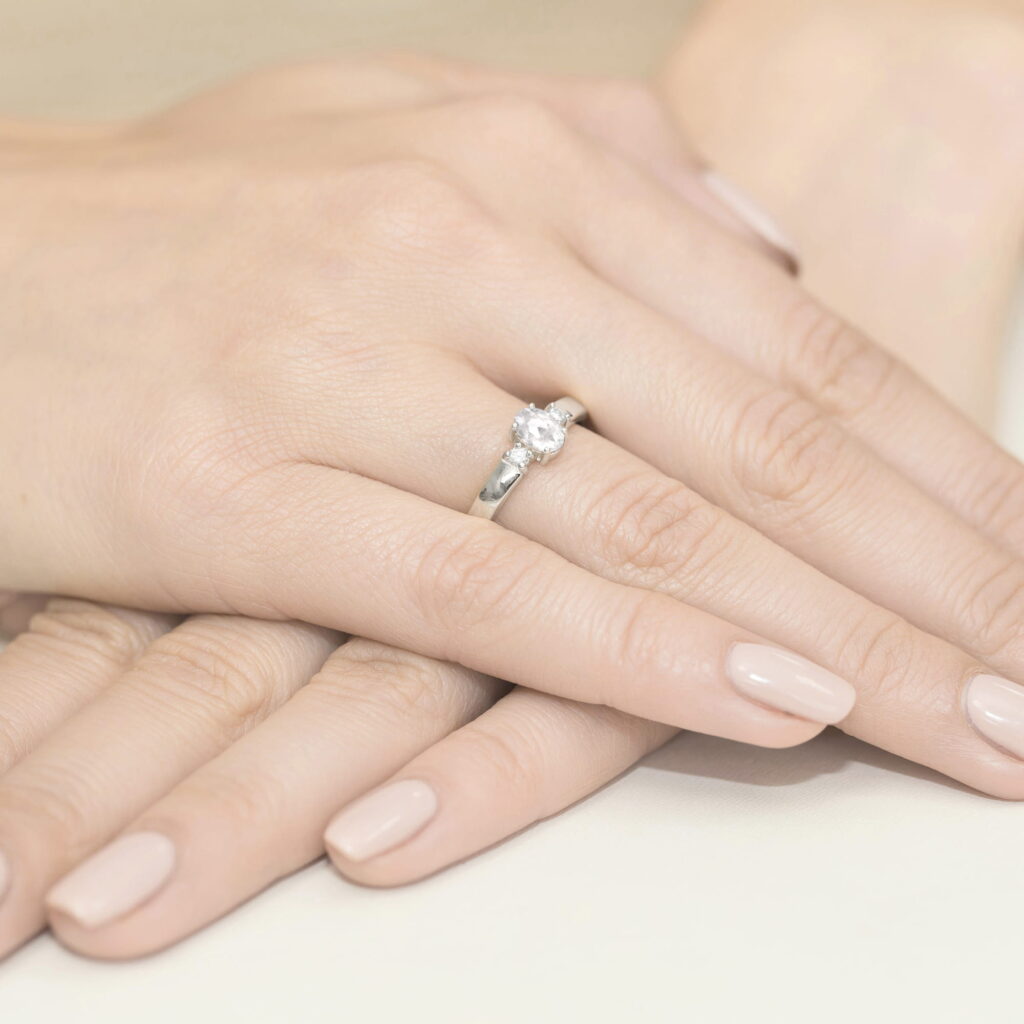 Na dłoni widoczny pierścionek z diamentem, otoczony delikatnymi, zadbanymi palcami z jasnym manicure.