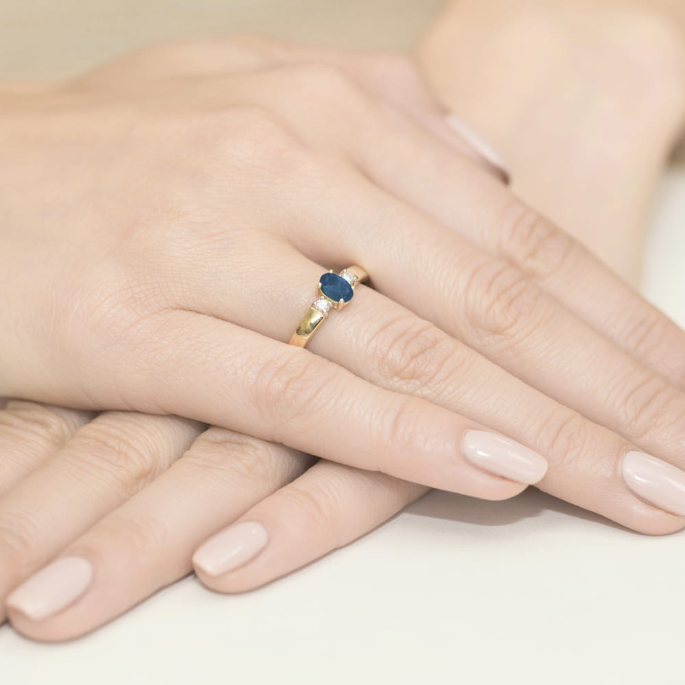 W0264.Z3.SAB.01_4 Złoty pierścionek z niebieskim kamieniem na palcu, dłonie z delikatnym manicure w jasnym kolorze.