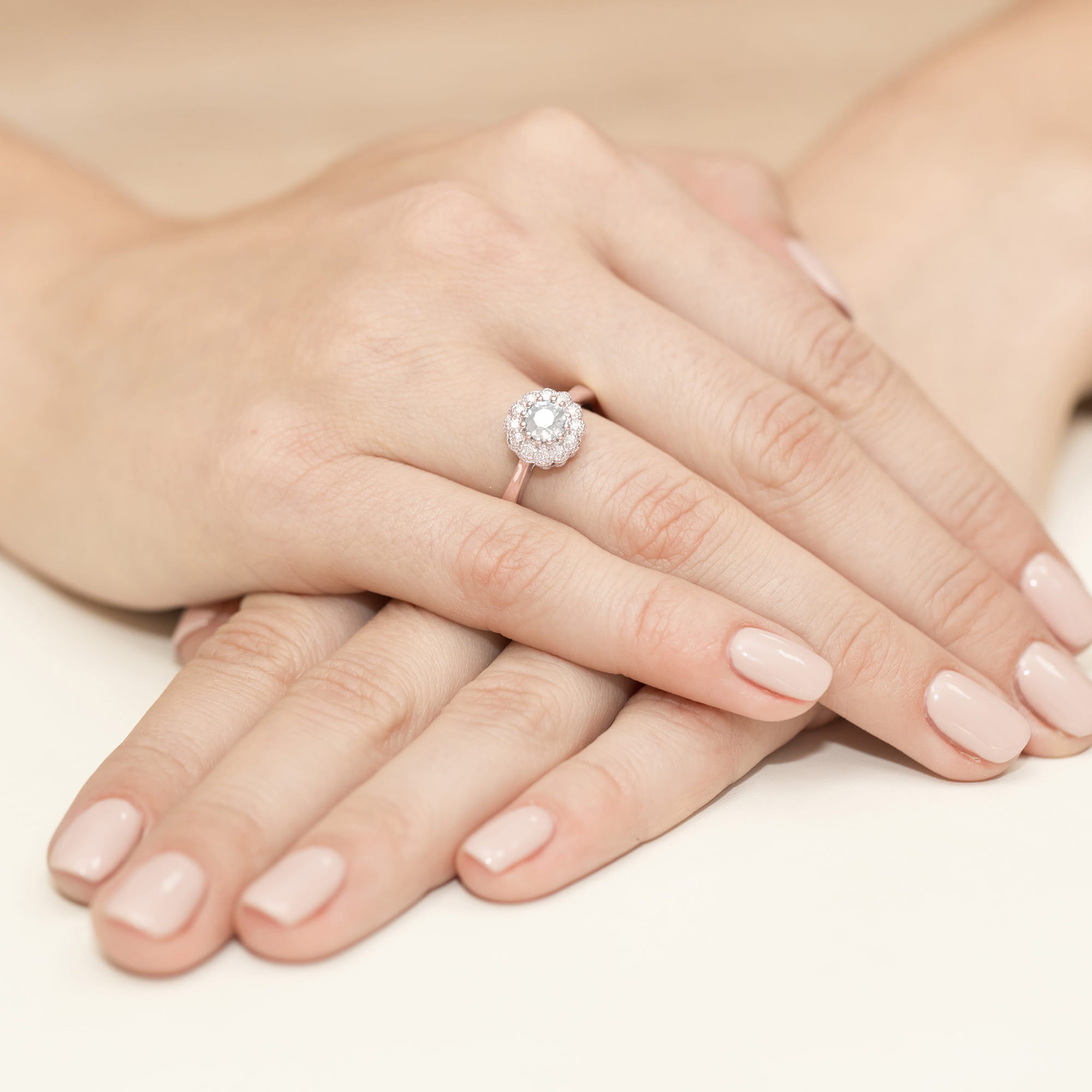 W0284.R3.LDW.01_4 Na dłoniach z delikatnym manicure znajduje się pierścionek z dużym, okrągłym diamentem otoczonym mniejszymi kamieniami.