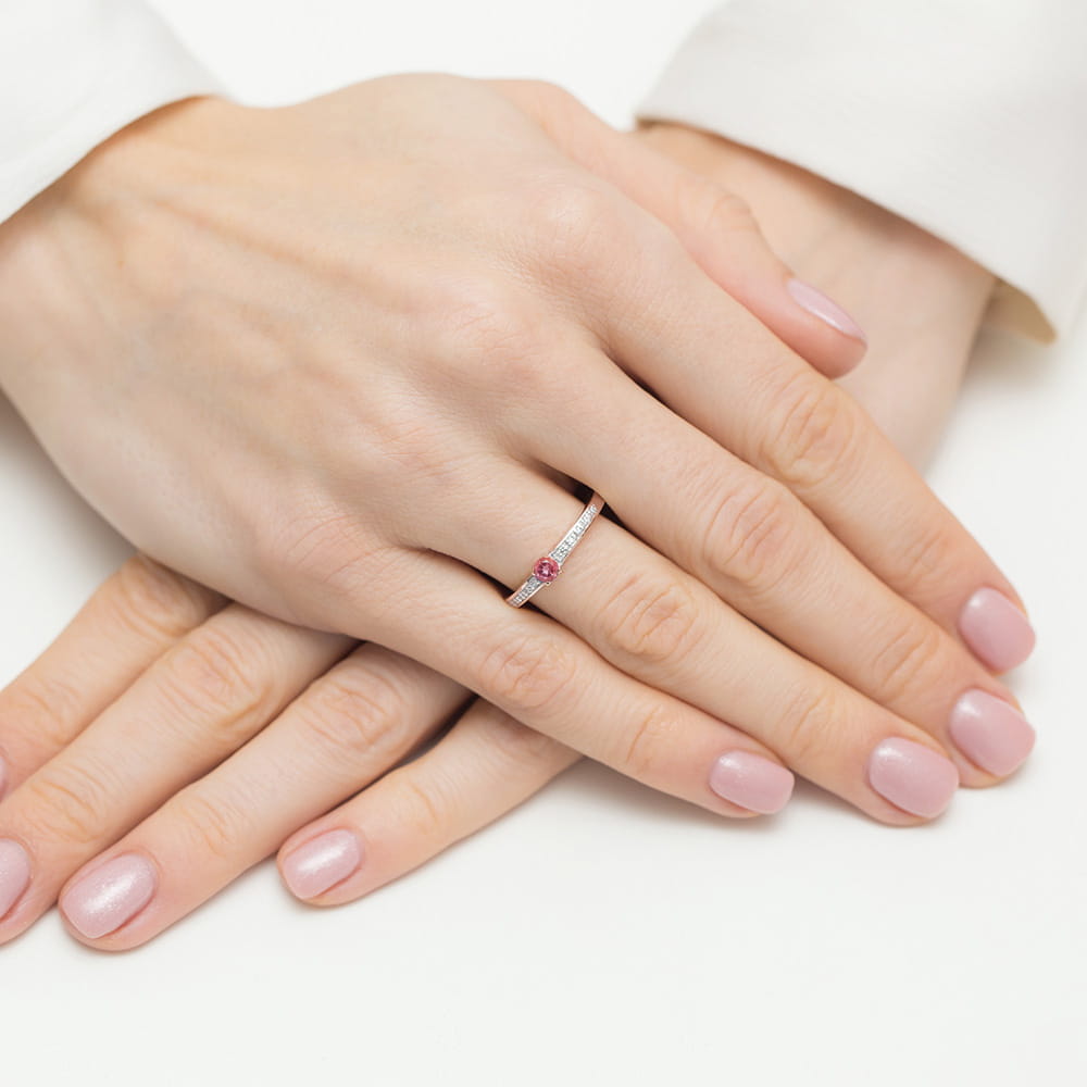 W0288.R3.TOP.01_4 Dwie ręce z delikatnym pierścionkiem z różowym kamieniem na palcu, z zadbanymi paznokciami w jasnym kolorze.