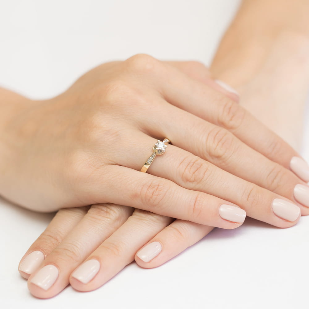 W0291.Z3.DIW.01_4 Na dłoni z delikatnym manicure znajduje się złoty pierścionek z dużym, błyszczącym kamieniem.