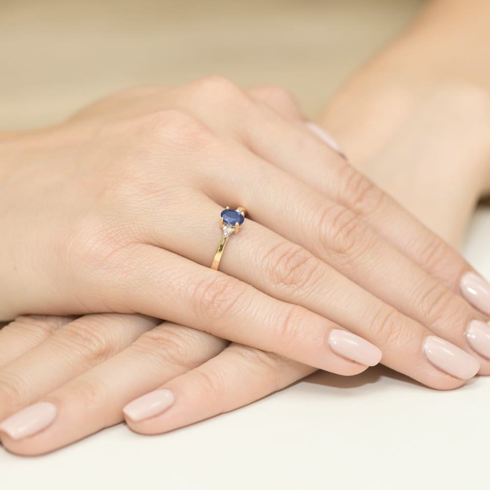 W0301.Z3.TAP.01_4 Złoty pierścionek z niebieskim kamieniem na palcu, dłonie z zadbanymi paznokciami w jasnym kolorze.