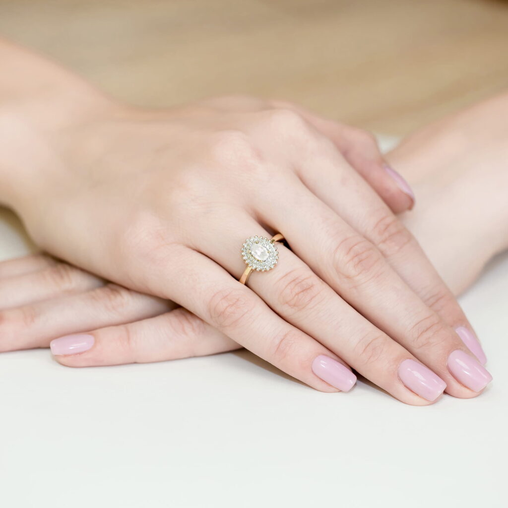 Dwie ręce z delikatnym manicure, jedna z pierścionkiem z okrągłym diamentem na złotej obrączce.