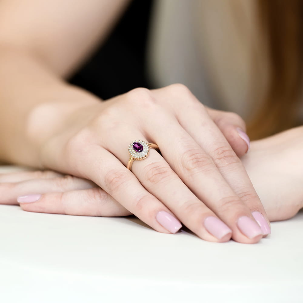 W0303.Z3.RHR.01_4 Złoty pierścionek z fioletowym kamieniem i diamentowym otoczeniem na dłoni z różowymi paznokciami.