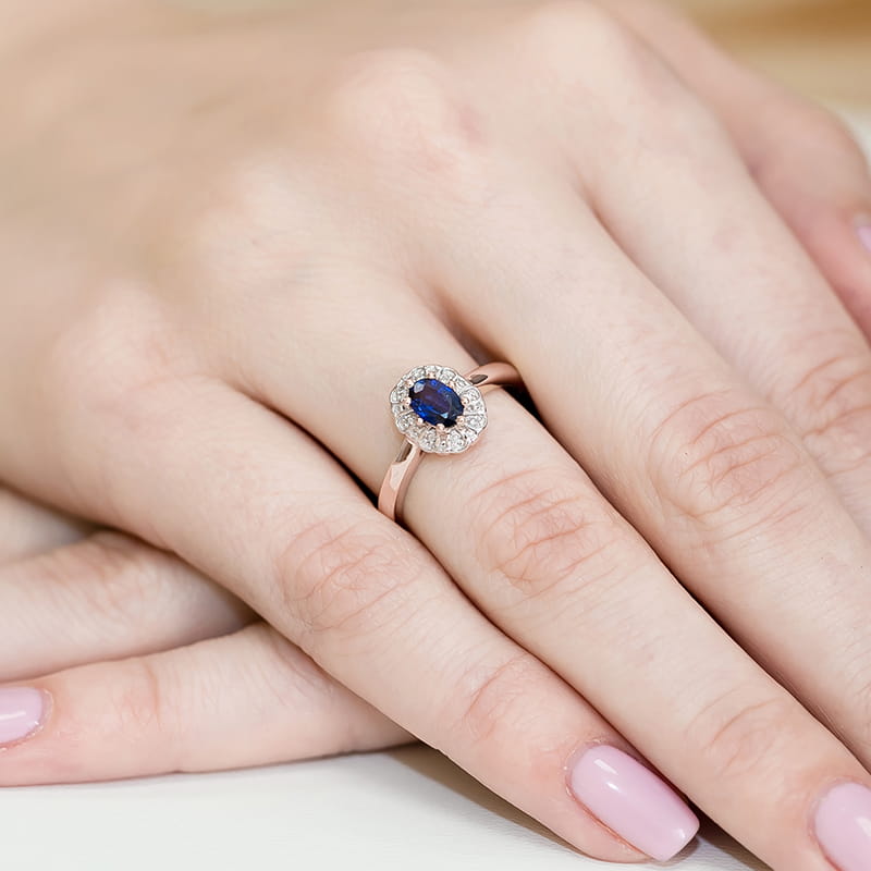 W0304.R3.SAB.01_4 Pierścionek z niebieskim kamieniem otoczony diamentami na palcu z różowym manicure.