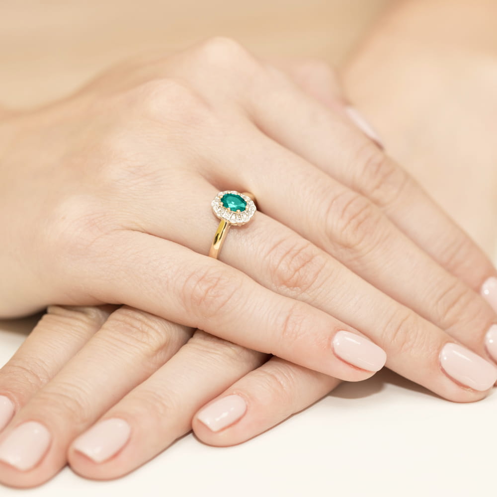 W0304.Z3.EMG.01_4 Złoty pierścionek z zielonym kamieniem otoczonym diamentami na dłoni z zadbanymi paznokciami.