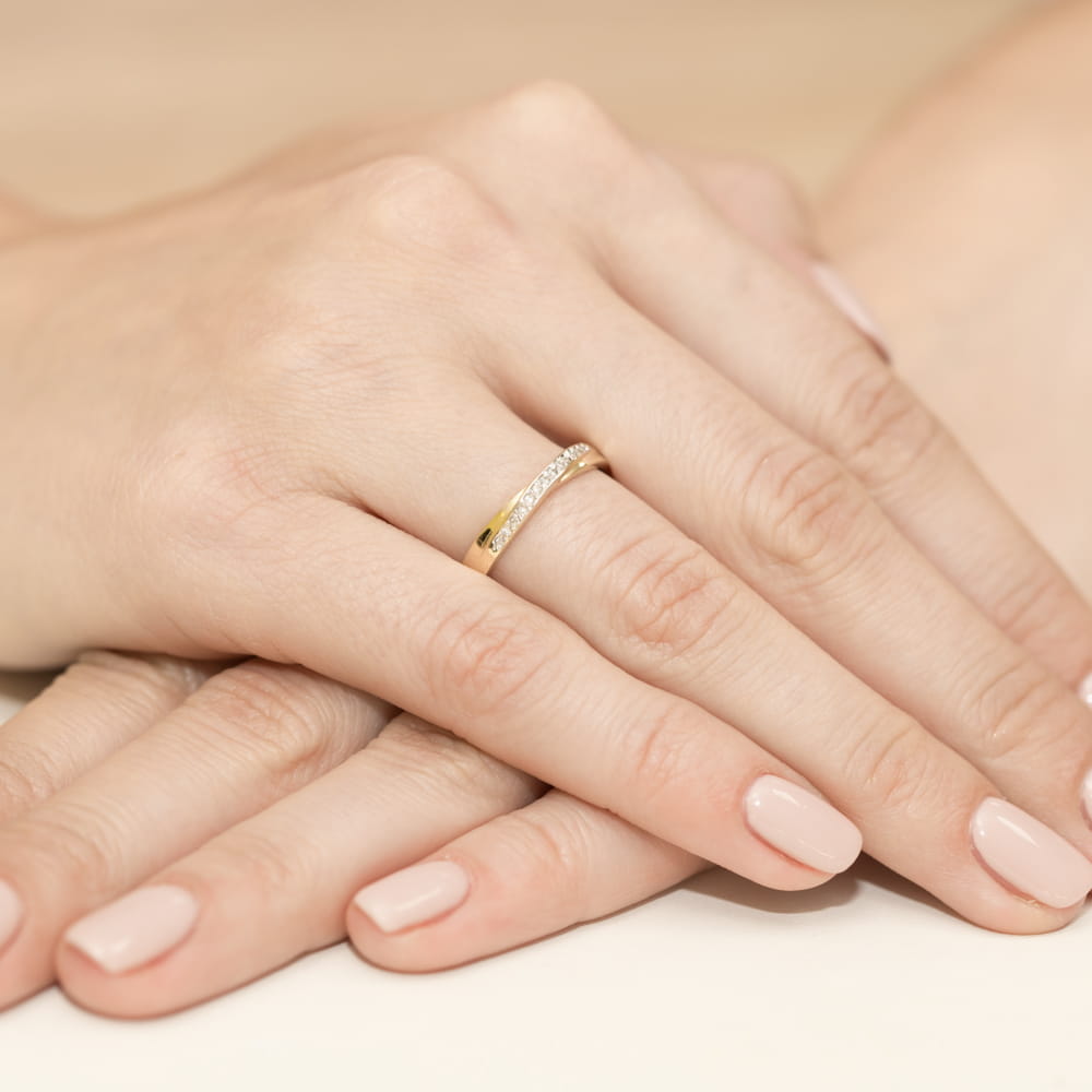 W0305.Z3.DIW.01_4 Dwie ręce z delikatnym złotym pierścionkiem ozdobionym diamentami, na tle jasnej powierzchni.