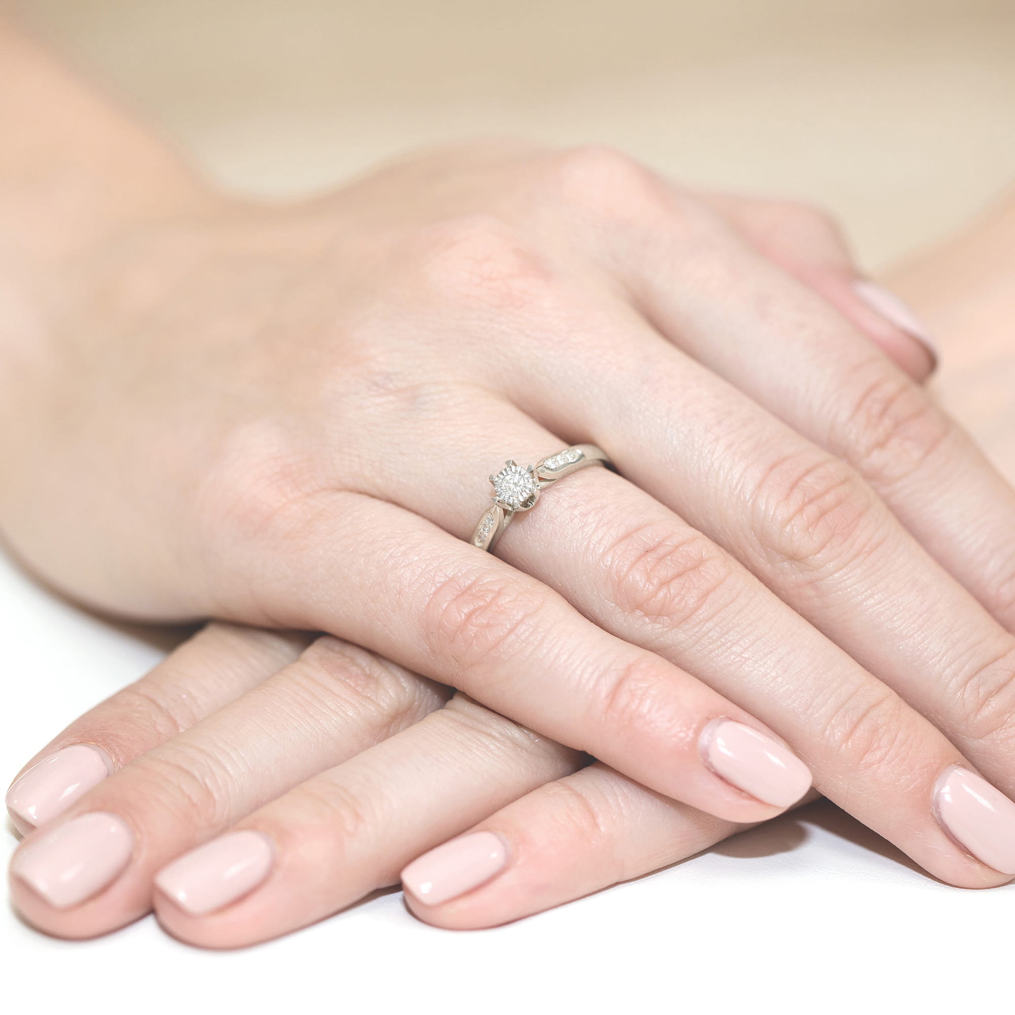 W0328.B3.DIW.01_4 Na dłoniach z delikatnym manicure znajduje się pierścionek z diamentem, otoczony subtelnym wzorem.