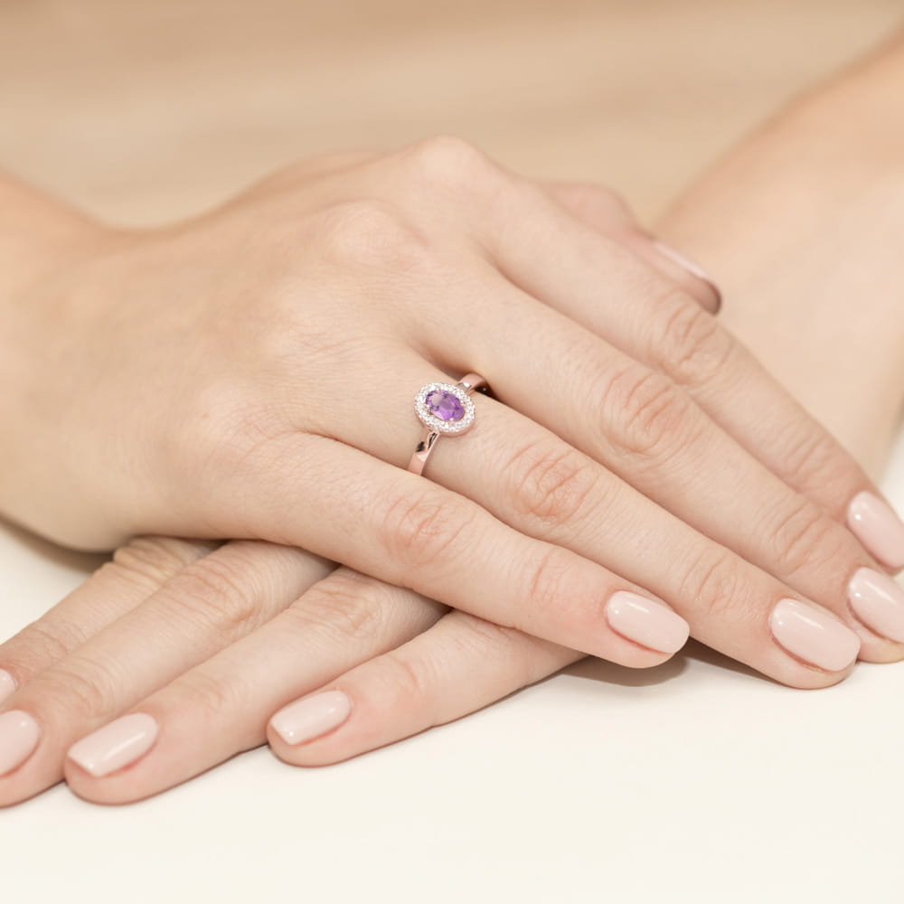 W0330.R3.AMV.01_4 Na dłoni z delikatnym manicure znajduje się pierścionek z fioletowym kamieniem otoczonym diamentami.