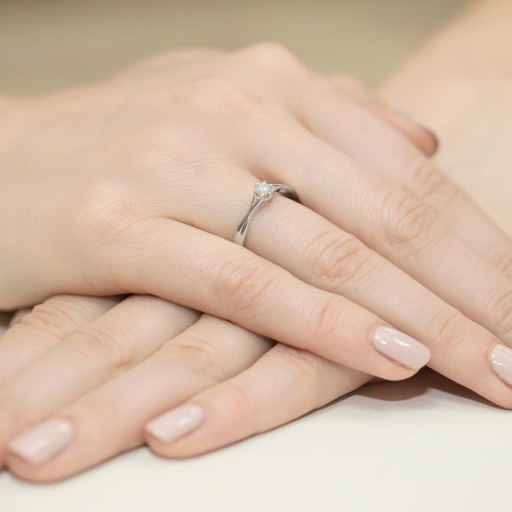 W0340.B3.DIW.02_4 Na dłoni widoczny jest srebrny pierścionek z diamentem, otoczony delikatnymi, zadbanymi dłońmi.