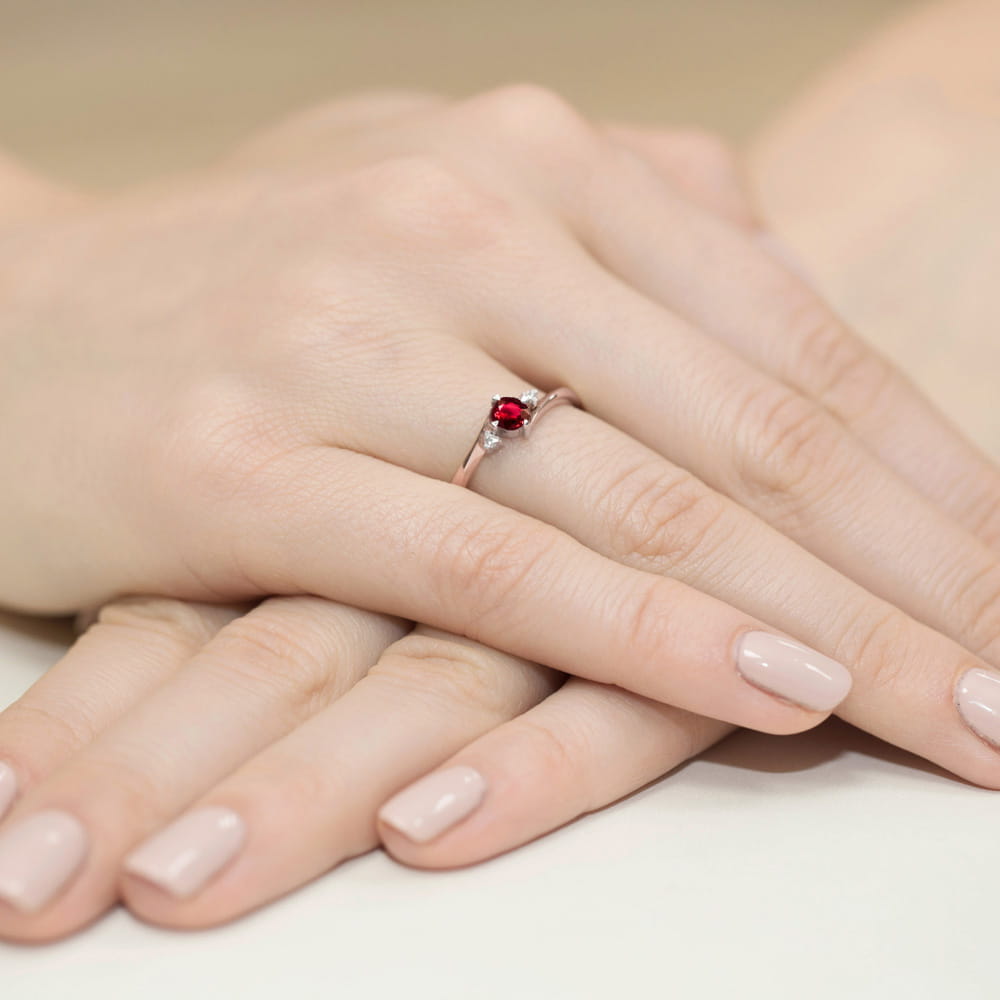 W0346.R3.RUR.02_4 Na dłoni znajduje się srebrny pierścionek z czerwonym kamieniem, otoczony delikatnymi palcami z jasnym manicure.