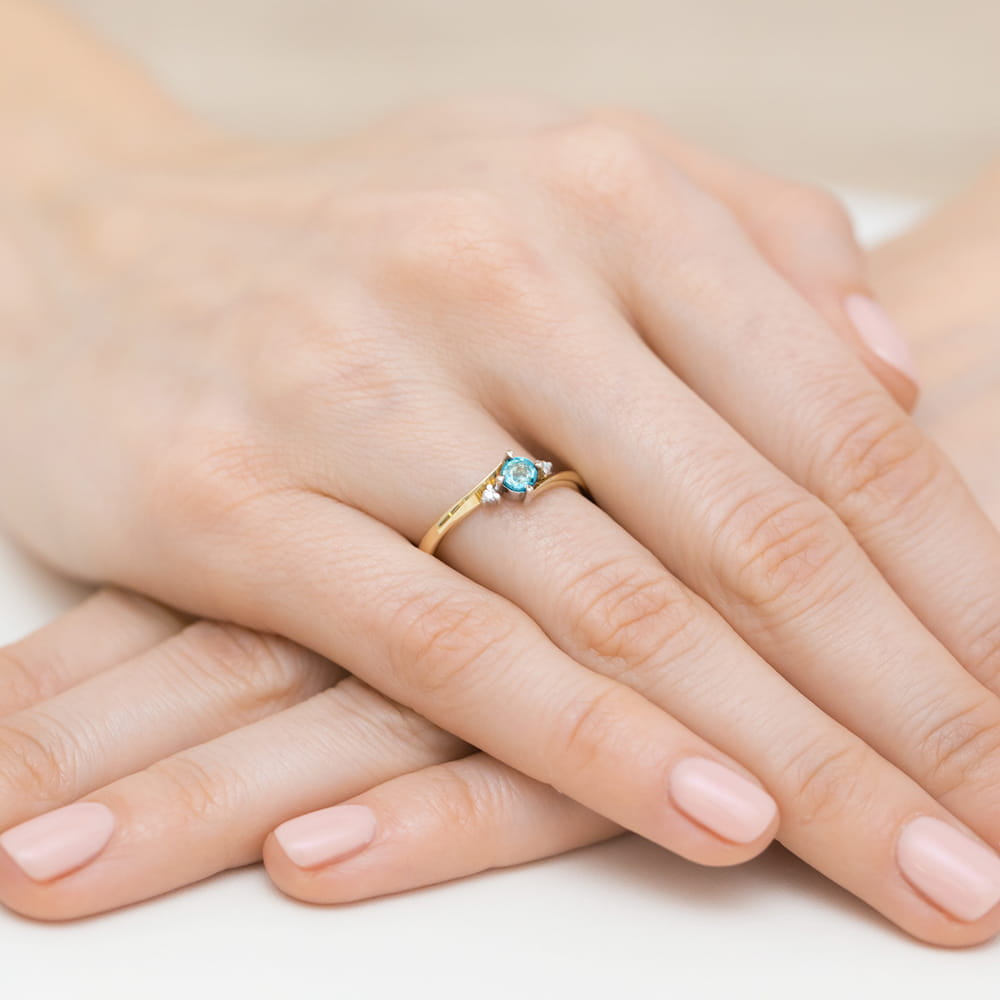 W0346.Z3.TOB.02_3 Złoty pierścionek z niebieskim kamieniem na palcu kobiety, dłonie z delikatnym manicure.