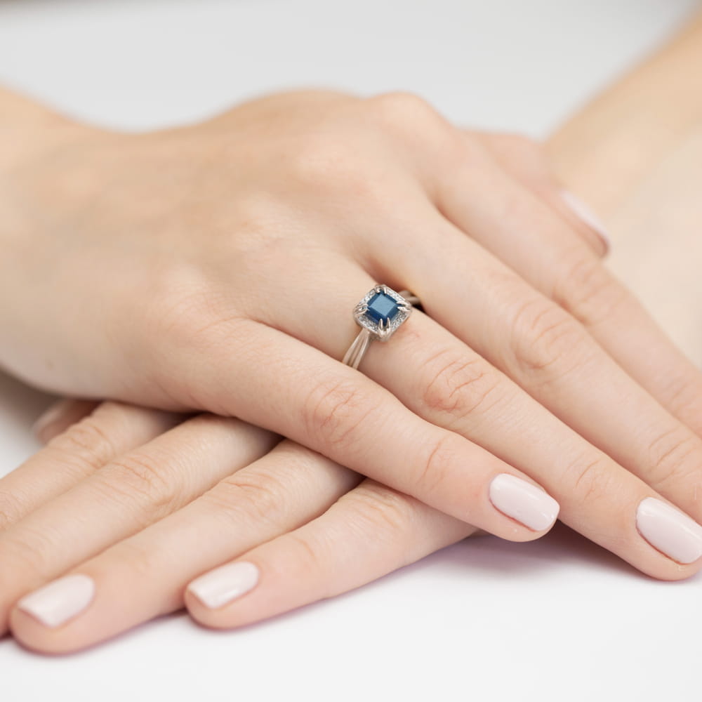 W0358.B3.SAB.01_4 Na dłoni z delikatnym manicure znajduje się pierścionek z niebieskim kamieniem szlachetnym.