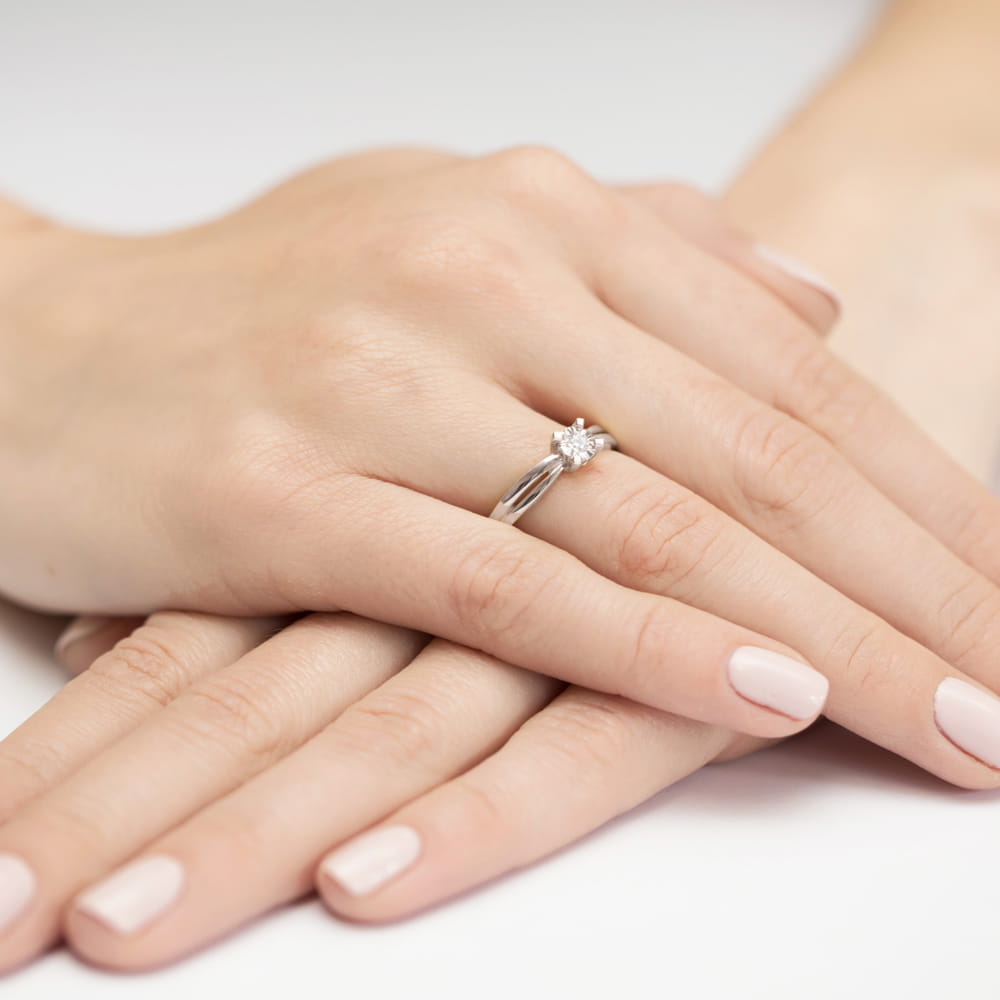 W0360.B3.DIW.01_4 Na dłoni widoczny pierścionek z diamentem, otoczony delikatnymi, zadbanymi palcami.