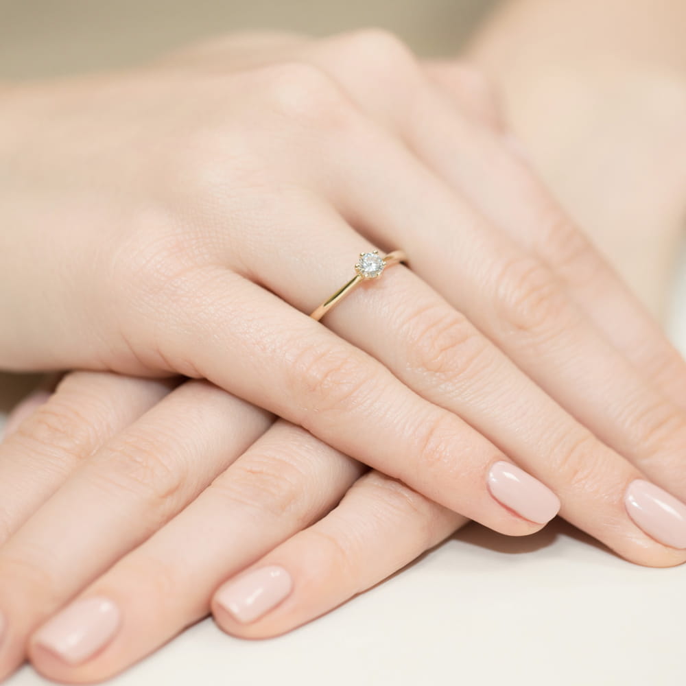 W0365.Z3.DIW.01_4 Złoty pierścionek z diamentem na palcu kobiety, dłonie z delikatnym manicure w odcieniu nude.