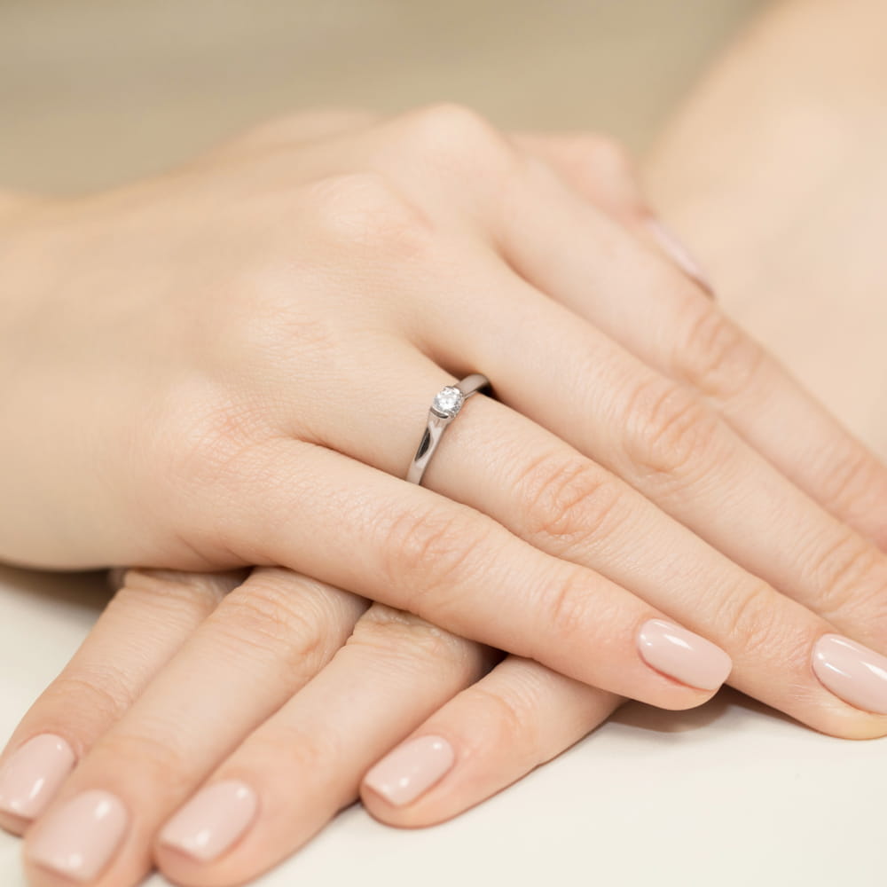 W0369.B3.DIW.01_4 Na dłoni widać srebrny pierścionek z diamentem, otoczony delikatnymi, zadbanymi paznokciami.