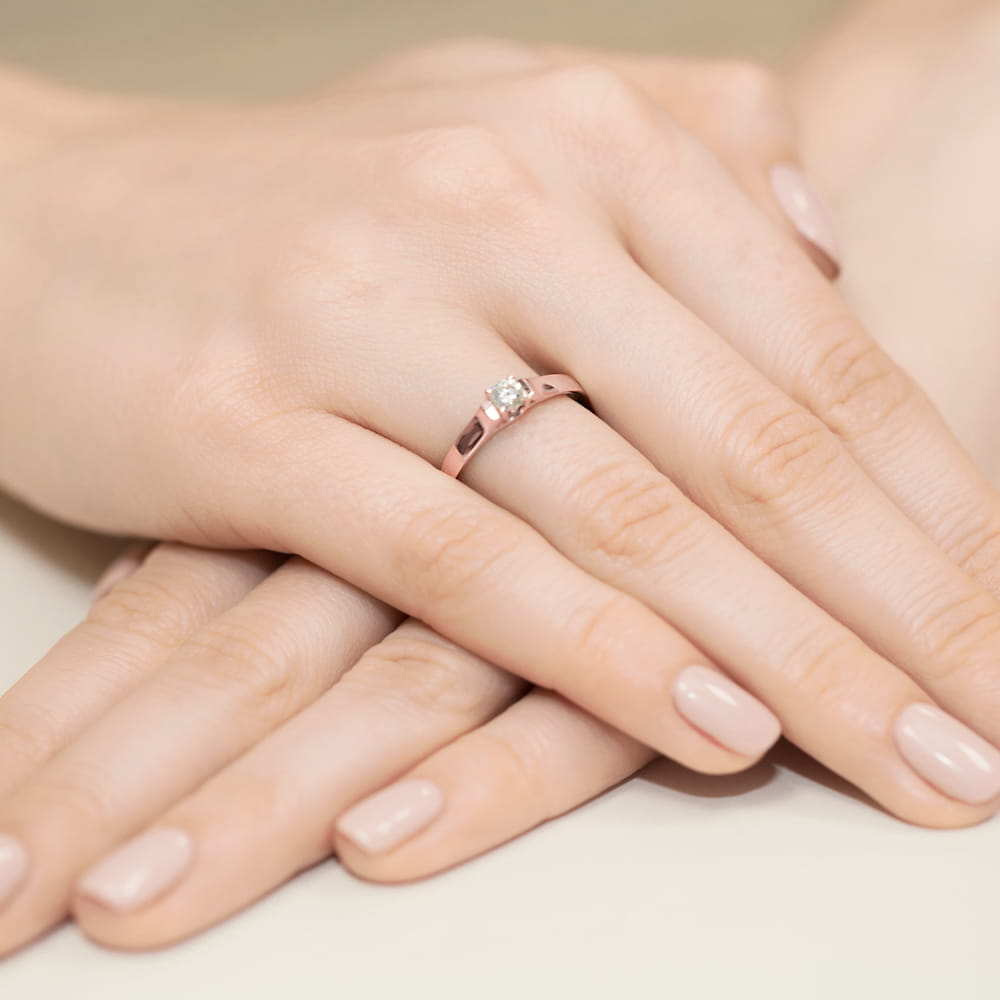 W0381.R3.DIW.01_4 Na dłoni widać różowe złote pierścionek z diamentem, otoczony zadbanymi, jasnymi paznokciami.