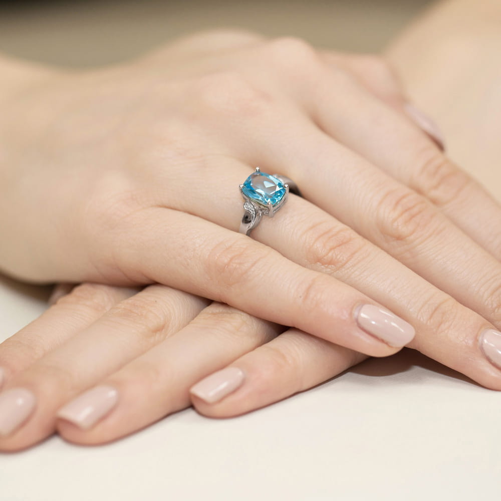 W0400.B3.TOB.01_4 Na dłoni widoczny srebrny pierścionek z dużym, niebieskim kamieniem, otoczony delikatnymi zdobieniami.