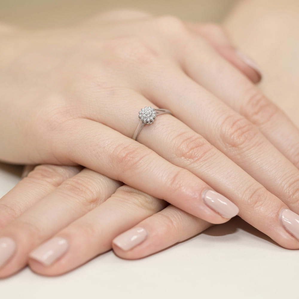 W0401.B3.DIW.01_4 Na dłoni widać pierścionek z diamentem, otoczony delikatnymi, jasnymi paznokciami.