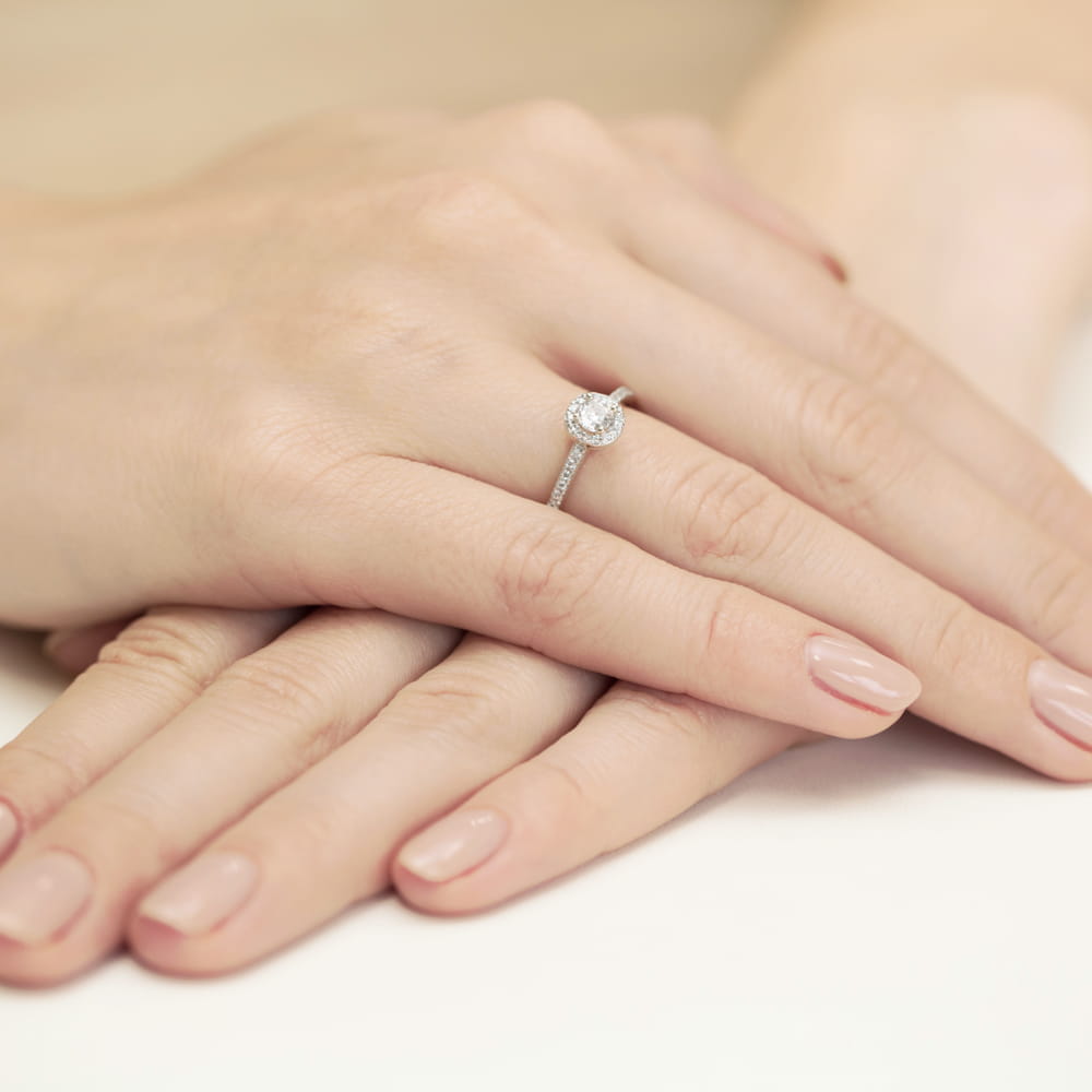 W0406.B3.LDW.01_4 Na dłoni widać pierścionek z okrągłym diamentem i delikatną obrączką, otoczony zadbanymi paznokciami.