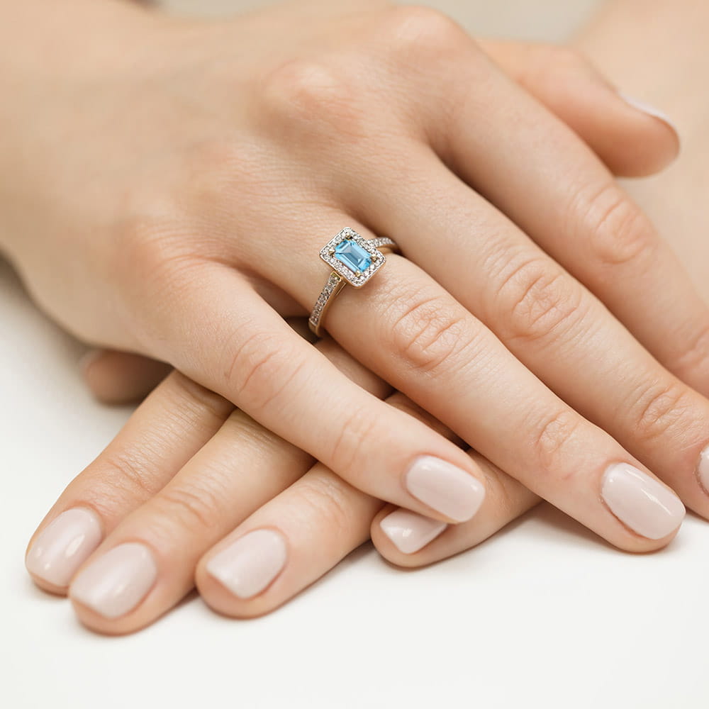 W0422.Z3.TOP.01_4 Pierścionek z niebieskim kamieniem w otoczeniu drobnych diamentów na palcu dłoni z zadbanymi paznokciami.