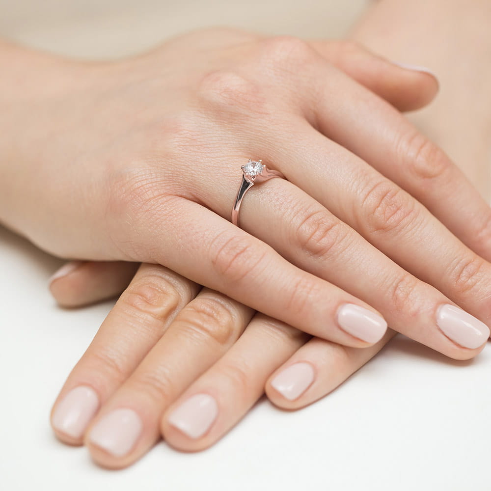W0423.R3.DIW.01_4 Na dłoni znajduje się delikatny pierścionek z małym diamentem, otoczony zadbanymi, jasnymi paznokciami.