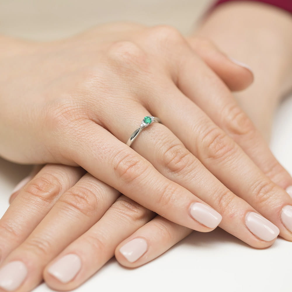 W0428.B3.EMG.01_4 Na dłoni widoczny srebrny pierścionek z zielonym kamieniem, otoczony delikatnymi, zadbanymi paznokciami.