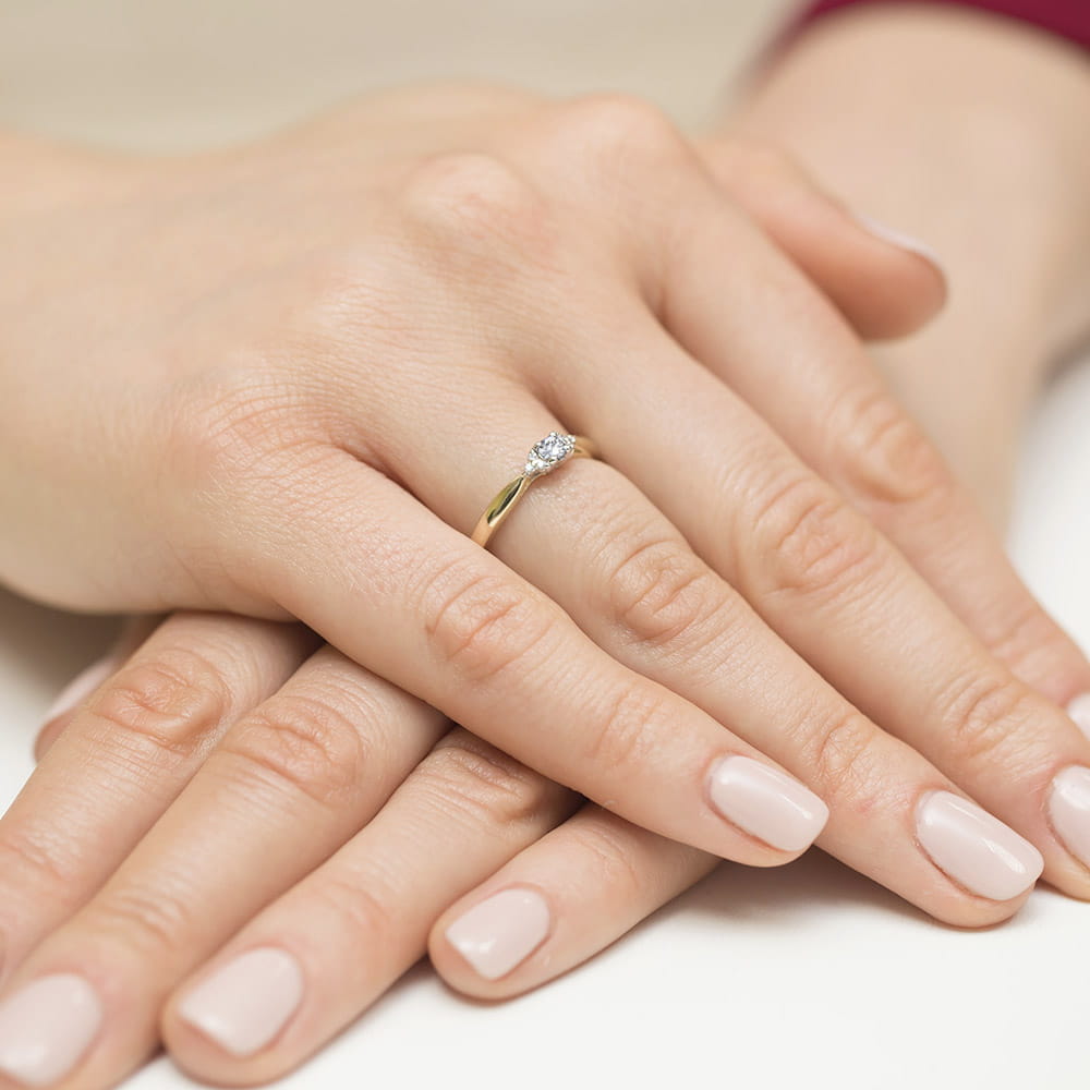 W0428.Z3.DIW.01_4 Złoty pierścionek z diamentem na palcu, dłonie z zadbanymi paznokciami ułożone na stole.
