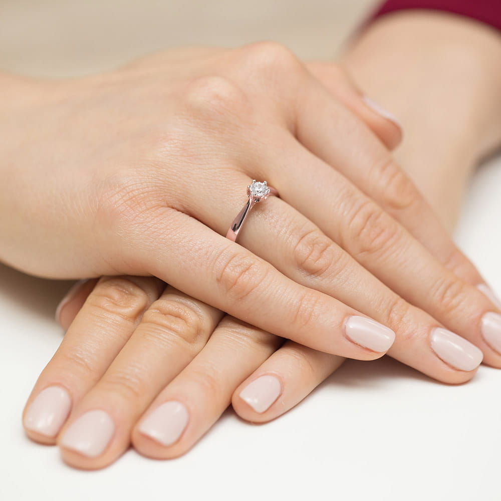 W0429.R3.DIW.01_4 Na dłoni z delikatnym manicure znajduje się pierścionek z diamentem, otoczony dwiema rękami.