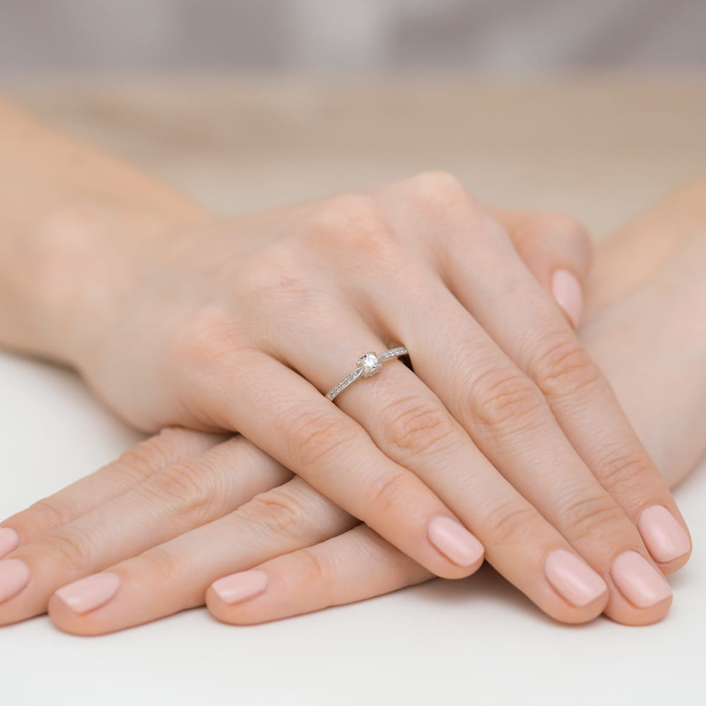 W0435.B3.LDW.01_4 Na dłoni z delikatnym manicure znajduje się pierścionek z diamentem, otoczony subtelnymi zdobieniami.