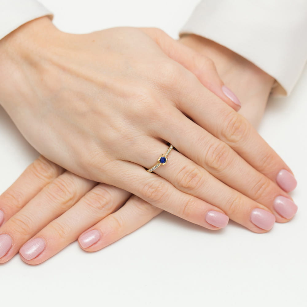 W0435.Z3.SAB.01_4 Na dłoni z różowym manicure znajduje się złoty pierścionek z niebieskim kamieniem.