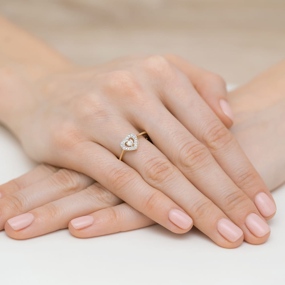 W0442.Z3.DIW.01_4 Dwie ręce z delikatnym pierścionkiem w kształcie serca, ozdobionym diamentami, na tle jasnej powierzchni.