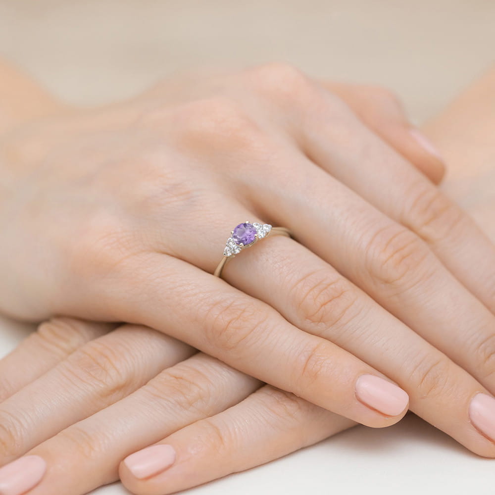 W0451.B3.AMV.01_4 Na dłoni widoczny złoty pierścionek z fioletowym kamieniem i diamentowymi zdobieniami.