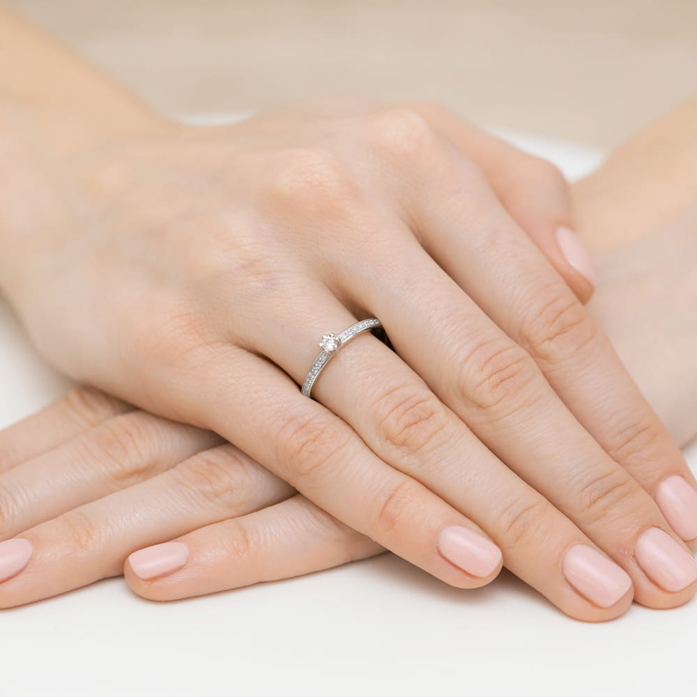 W0459.B3.DIW.01_4 Na dłoni z delikatnym manicure znajduje się pierścionek z diamentem i małymi kamieniami.