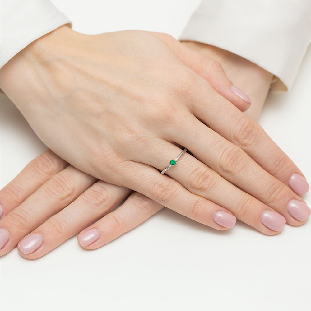 W0464.B3.EMG.01_4 Dwie ręce z delikatnym pierścionkiem z zielonym kamieniem na palcu, z zadbanymi paznokciami w jasnym kolorze.
