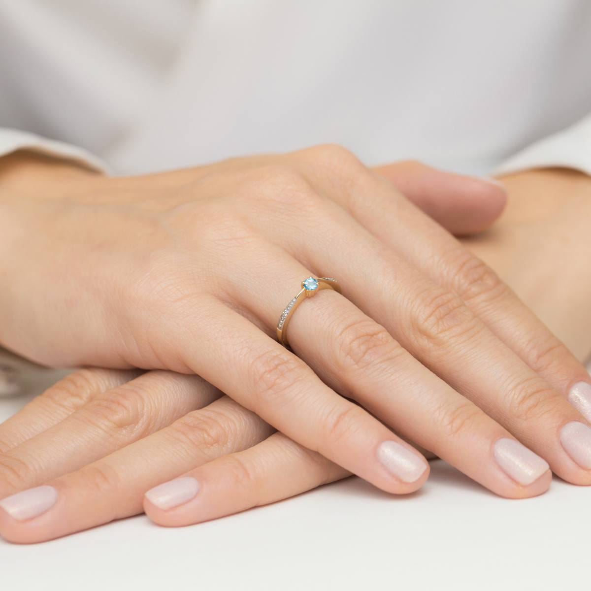 W0465.Z3.TOB.01_4 Złoty pierścionek z niebieskim kamieniem i diamentami na delikatnej dłoni z zadbanymi paznokciami.