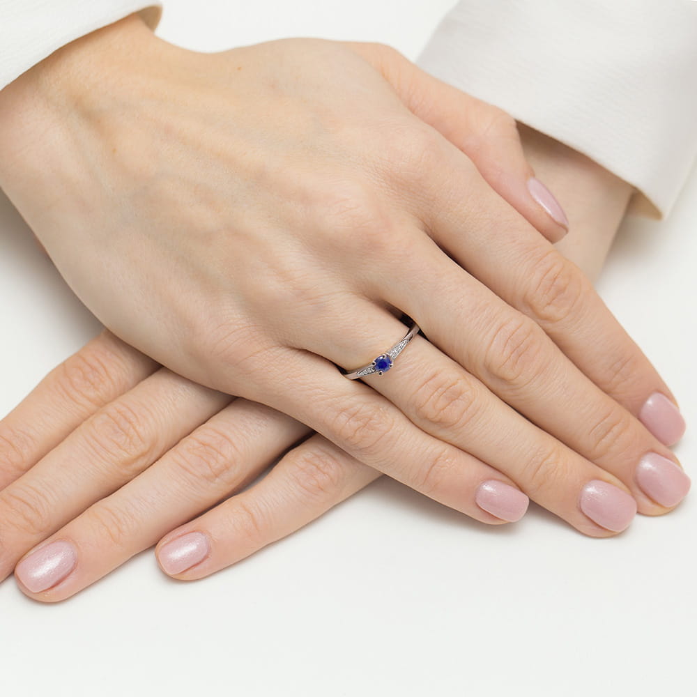 W0466.B3.SAB.01_4 Dwie ręce z delikatnym pierścionkiem z niebieskim kamieniem, paznokcie w jasnym kolorze.