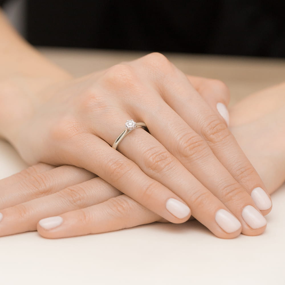 W0468.B3.DIW.01_4 Na dłoni znajduje się delikatny pierścionek z diamentem, otoczony gładkimi, zadbanymi paznokciami.