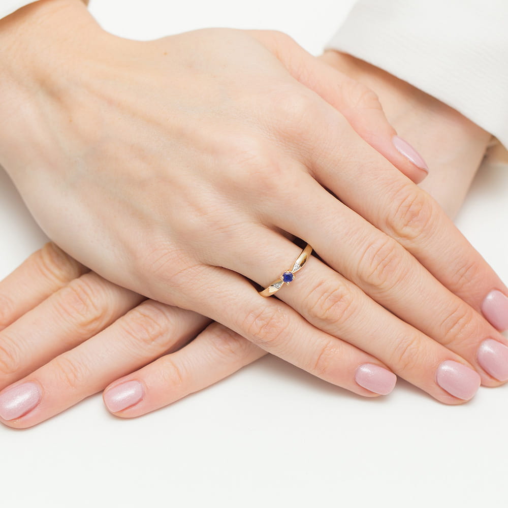 W0469.Z3.SAB.01_4 Dwie ręce z delikatnym pierścionkiem na palcu, z niebieskim kamieniem, na jasnym tle.