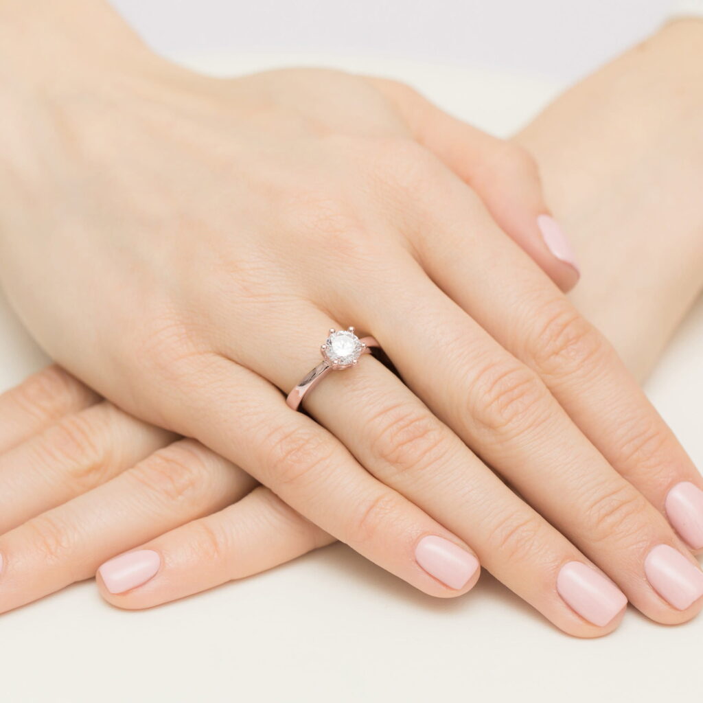 Na dłoni z różowym manicure znajduje się pierścionek z dużym, okrągłym diamentem.