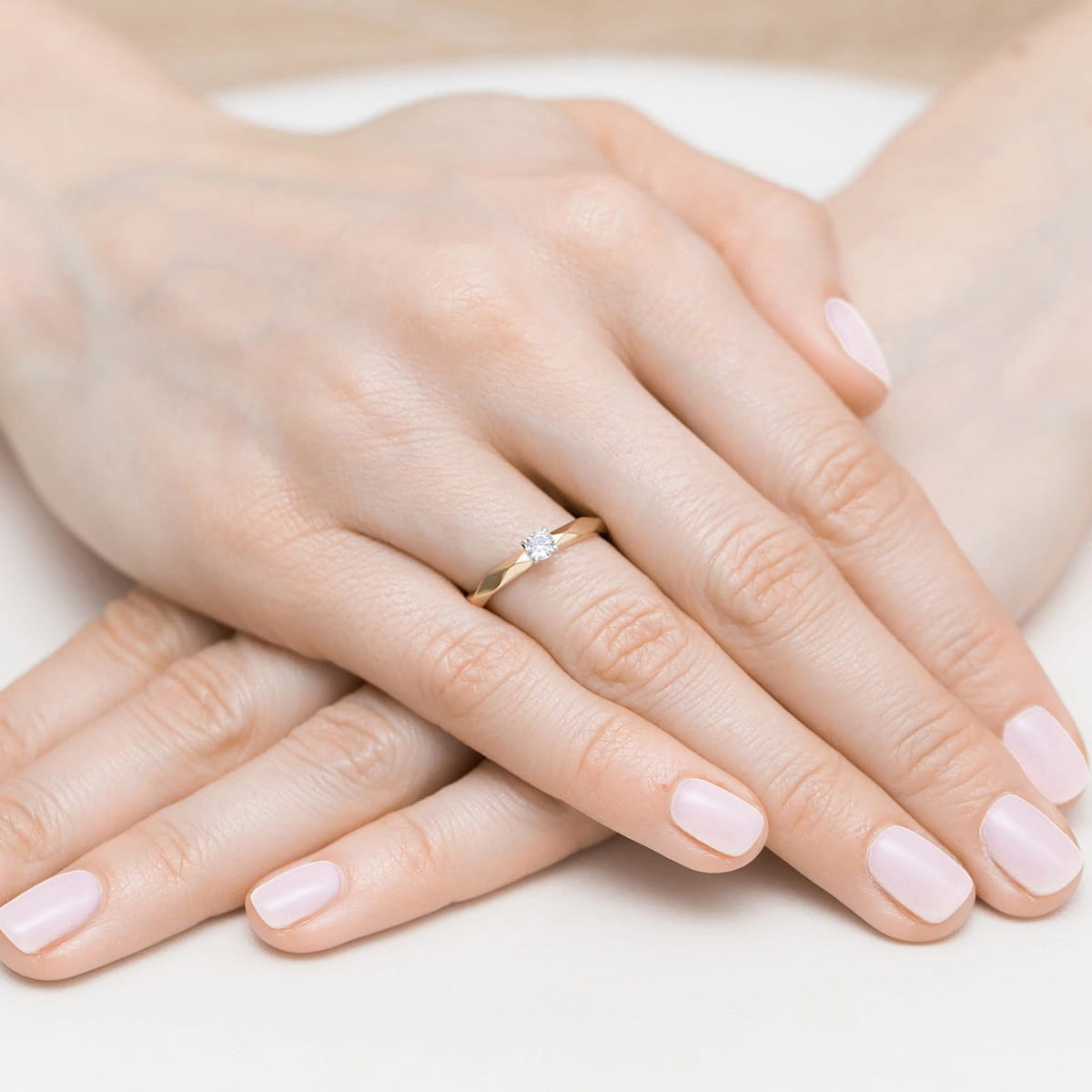 W0484.Z3.DIW.01_4 Dwie ręce z delikatnym pierścionkiem zaręczynowym na palcu, z różowymi paznokciami.