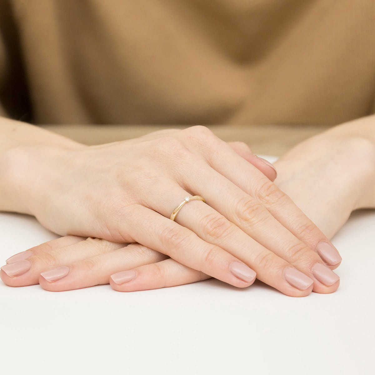 W0501.Z3.DIW.01_4 Dwie ręce z delikatnym złotym pierścionkiem, złożone na stole, z zadbanymi paznokciami w neutralnym kolorze.